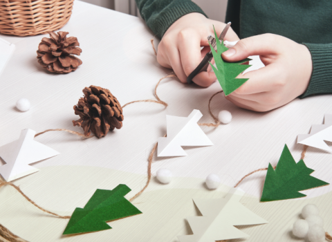 Child's hands cutting trees over table with paper and pinecones