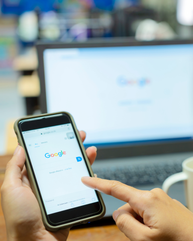 Hand holding device with Google onscreen in front of laptop with Google onscreen