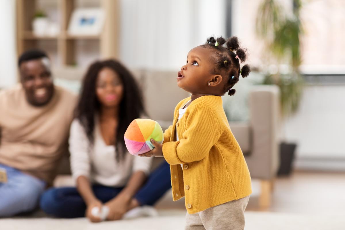 child age 12-18 months standing holding a ball while parents watch